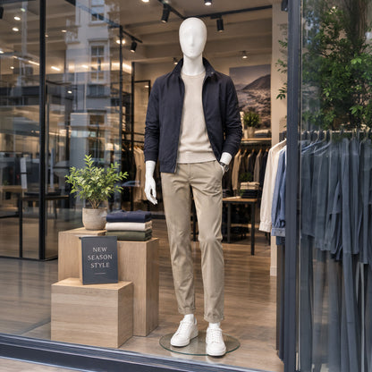 A mannequin dressed in a dark navy jacket, beige sweater, beige trousers, and white sneakers is displayed in a clothing store window display.