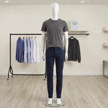 A mannequin in a minimalist clothing store setting, wearing a dark gray crewneck t-shirt and navy blue jeans. In the background, a clothing rack holds light blue and white shirts, and a black hoodie hangs on a wall rack next to a shelf.